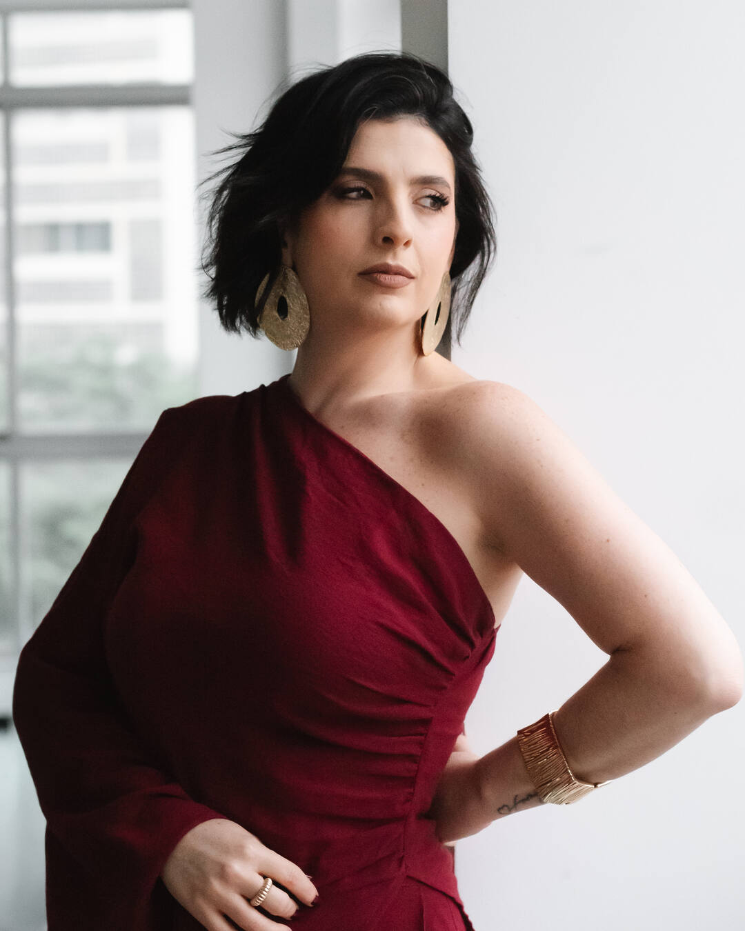 Bella Patera, de vestido bordô de um ombro só e brincos dourados, em frente a uma janela